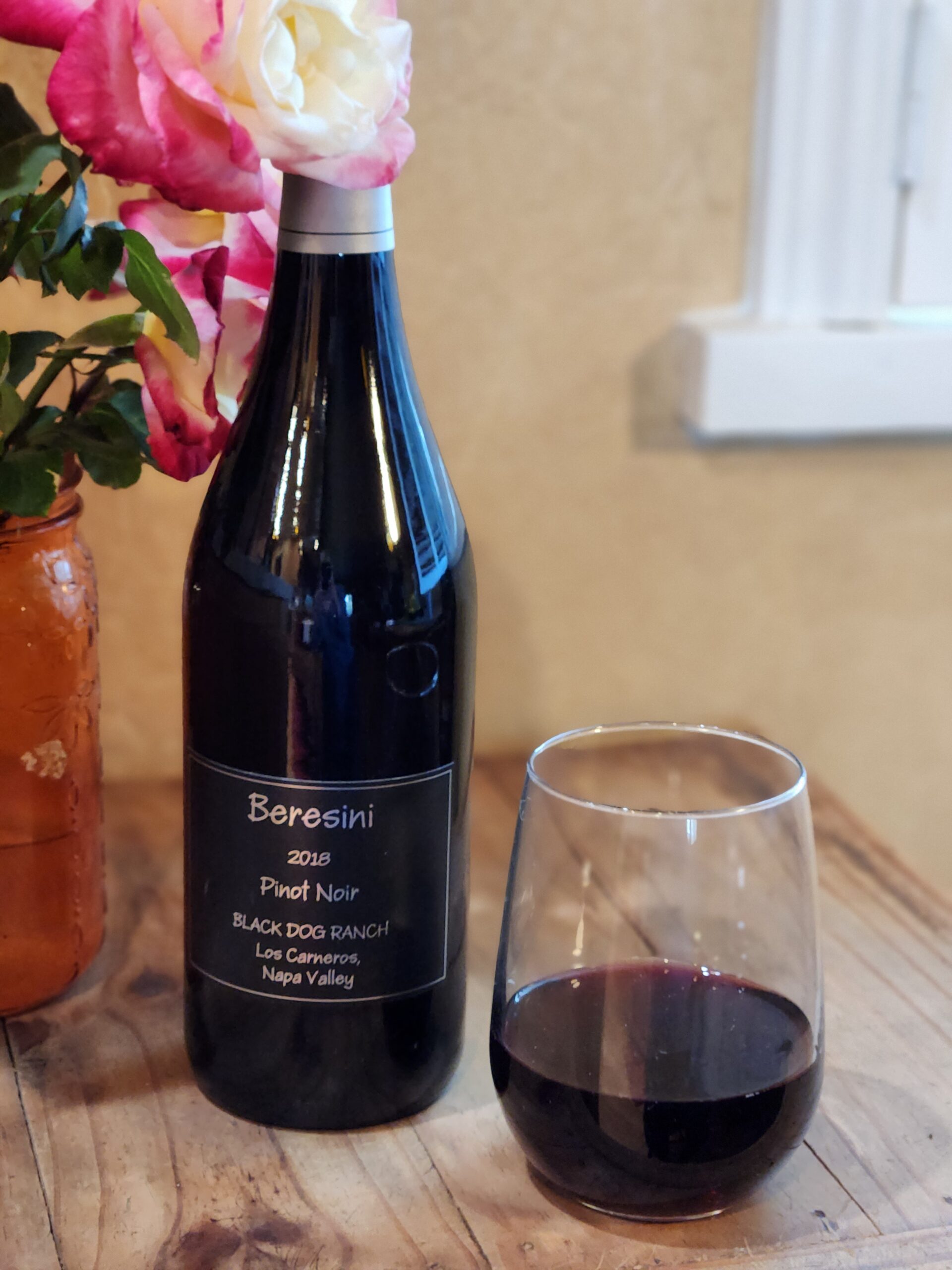 Bottle of 2019 Pinot Noir on a picnic table next to a vase of flowers and glass of wine